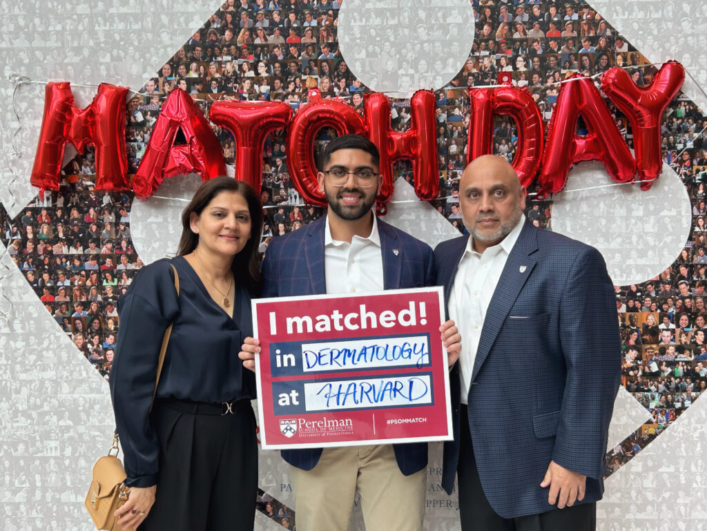 Photograph of a man in his 30s, he stands in front of a balloon sign that reads "Match Day" and he is holding a sign that says "I matched! in Dermatology at Harvard". He is flanked on either side by middle-aged man and woman.