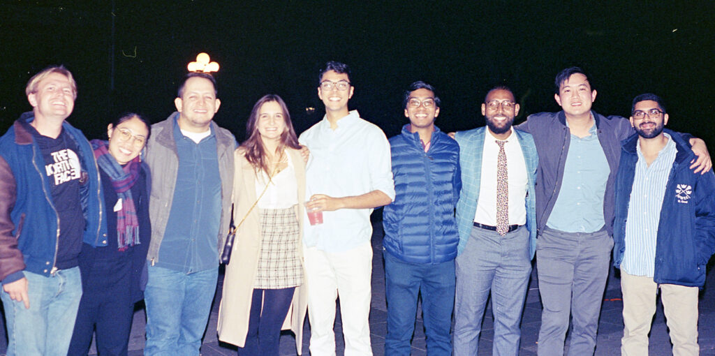 Photograph of a group of nine people, standing with their arms around each other. They are all in their 20s and have diverse heritages, they are all smiling. 