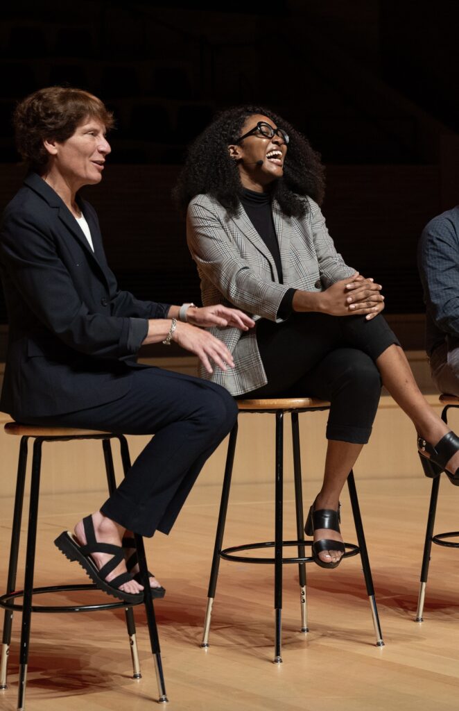 Photograph of a woman in her 20s with dark-medium skin tone and black curly hair, she is wearing black pants, black turtleneck and a grey tartan blazer. She is seated on a stool on a stage, with people on each side, she has a microphone on and is laughing. 