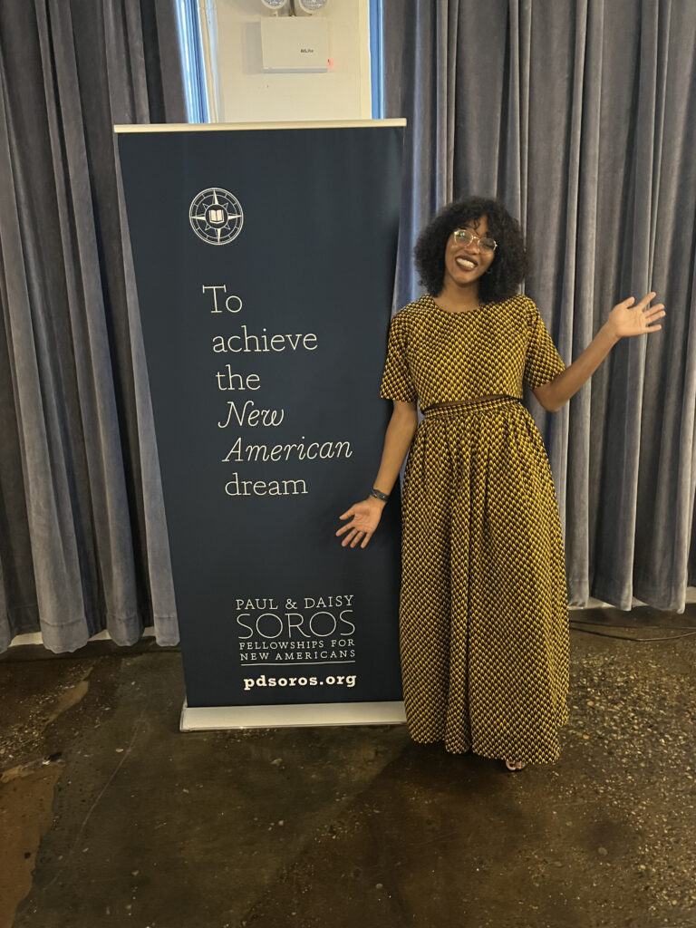 Photograph of a woman in her 20s with dark-medium skin tone and black shoulder length curly hair, she is wearing a matching top and skirt in black and yellow checkers. She is standing next to a banner that says "To achieve the New American dream" with the Paul & Daisy Soros logo at the bottom.