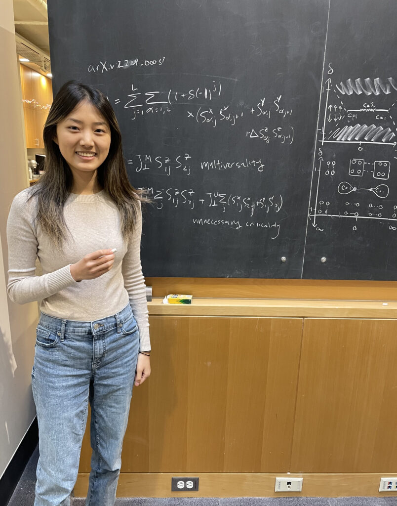 Photograph of a woman in her 20s standing in front of a chalkboard full of equations. She is smiling.