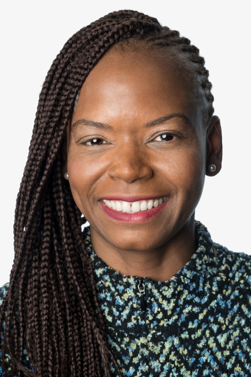 Headshot of a woman in her 40s with dark-medium skin tone and long braided black hair, pulled back on her left side. 