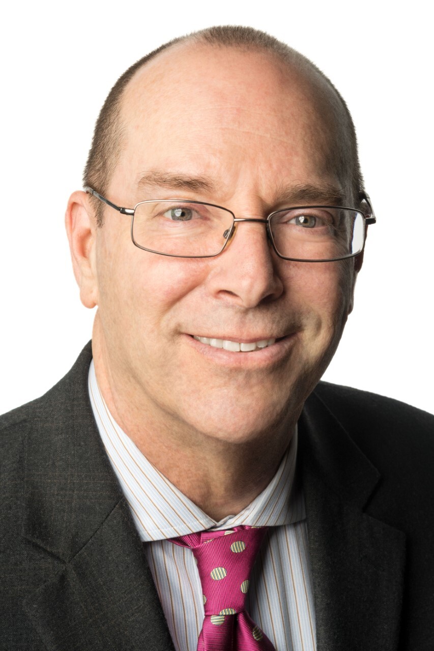 Headshot of a man in his 50s with light skin tone and brown balding hair. He is wearing a black suit and white button up and has on thin rimmed rectangle glasses. He is smiling at the camera. 