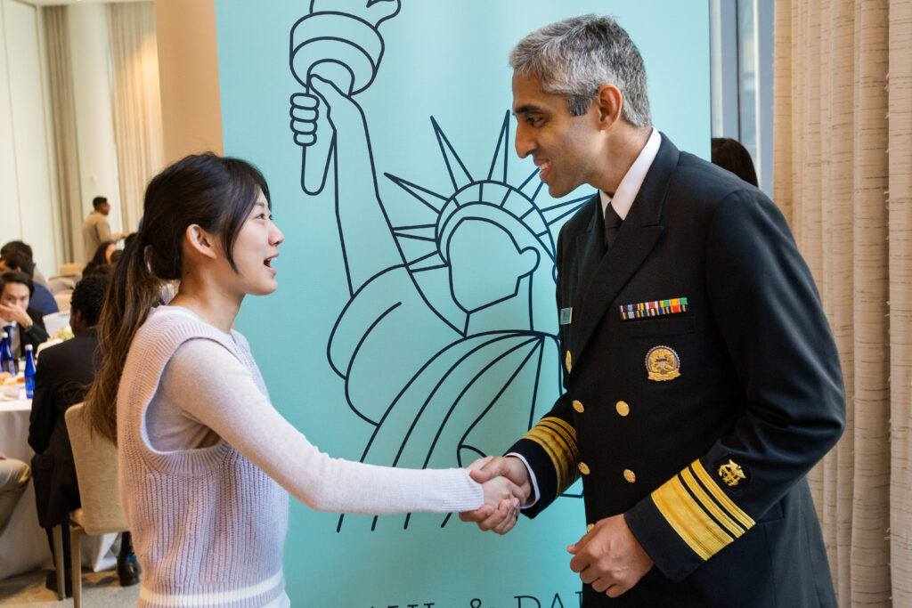 Photograph of a woman in her 20s wearing a light purple sweater, she is looking at and shaking hands with a man in his 40s wearing a military uniform. 