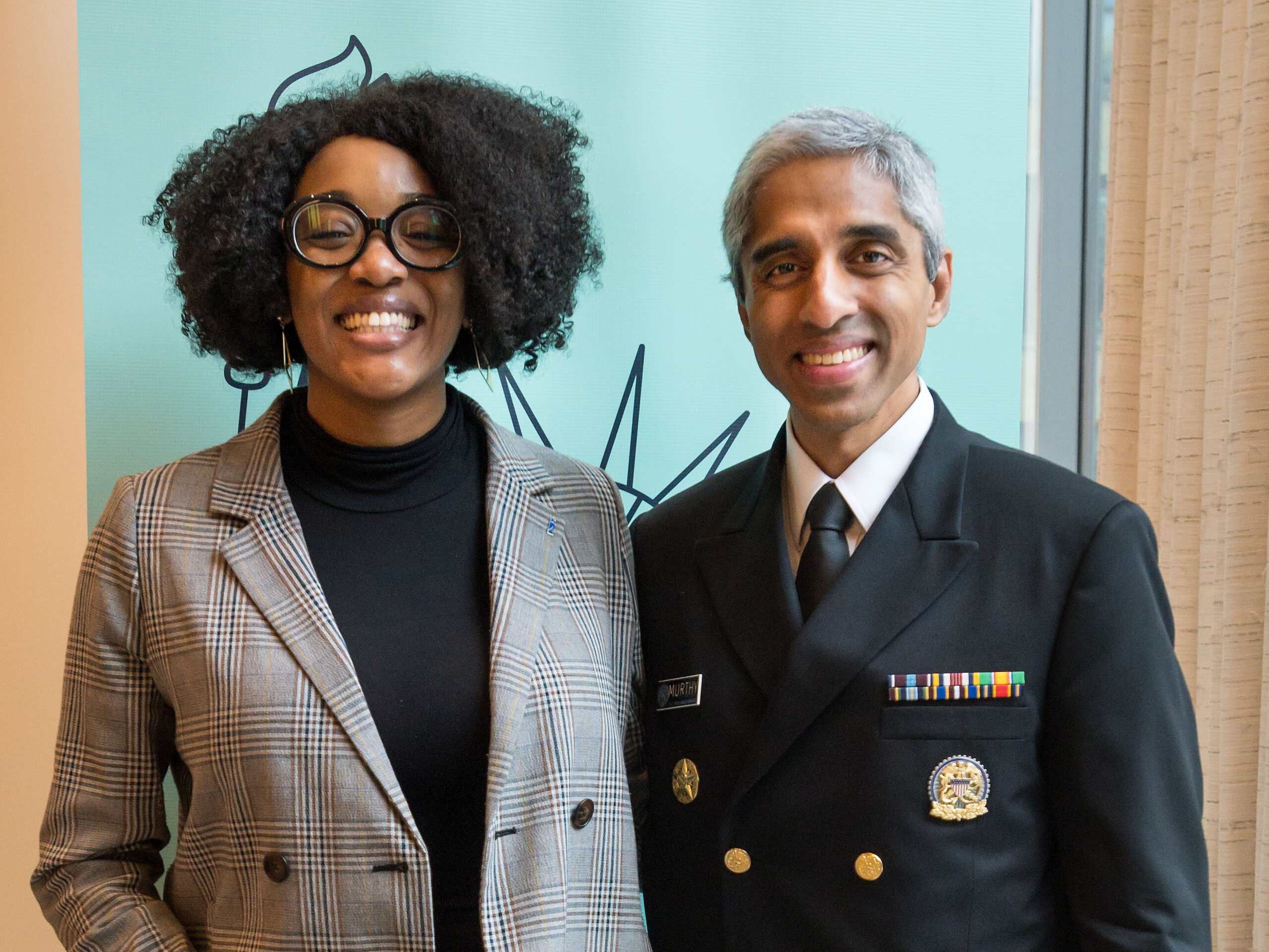 Photograph of a woman in her 20s with dark-medium skin tone and chin length curly black hair, wearing a black turtleneck and grey tartan blazer, smiling at the camera, She is standing next to a man in his 40s wearing a military uniform. 