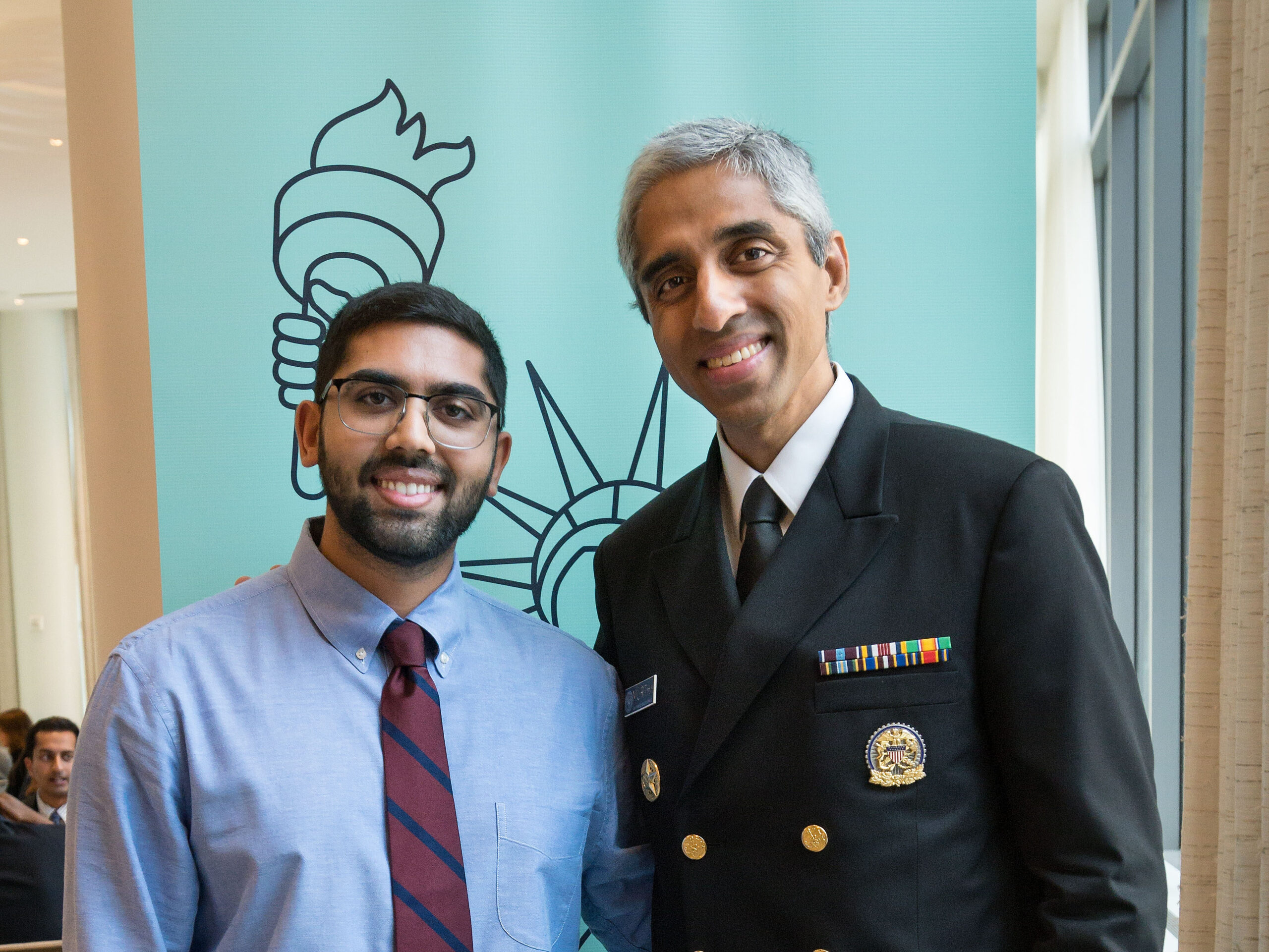 Photograph of a man in his 20s with medium skin tone, short black hair, and short beard. He is wearing a blue button up shirt and maroon tie with large thin rimmed glasses. He stands next to a man in his 40s wearing a military uniform. 