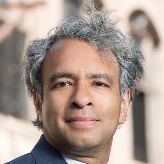Headshot of a man in his 40s who has heritage from India with light-medium skin tone and salt and pepper tousled hair. What he is wearing can barely be seen, a navy suit collar and white button up are peeking out. He is looking at the camera. 