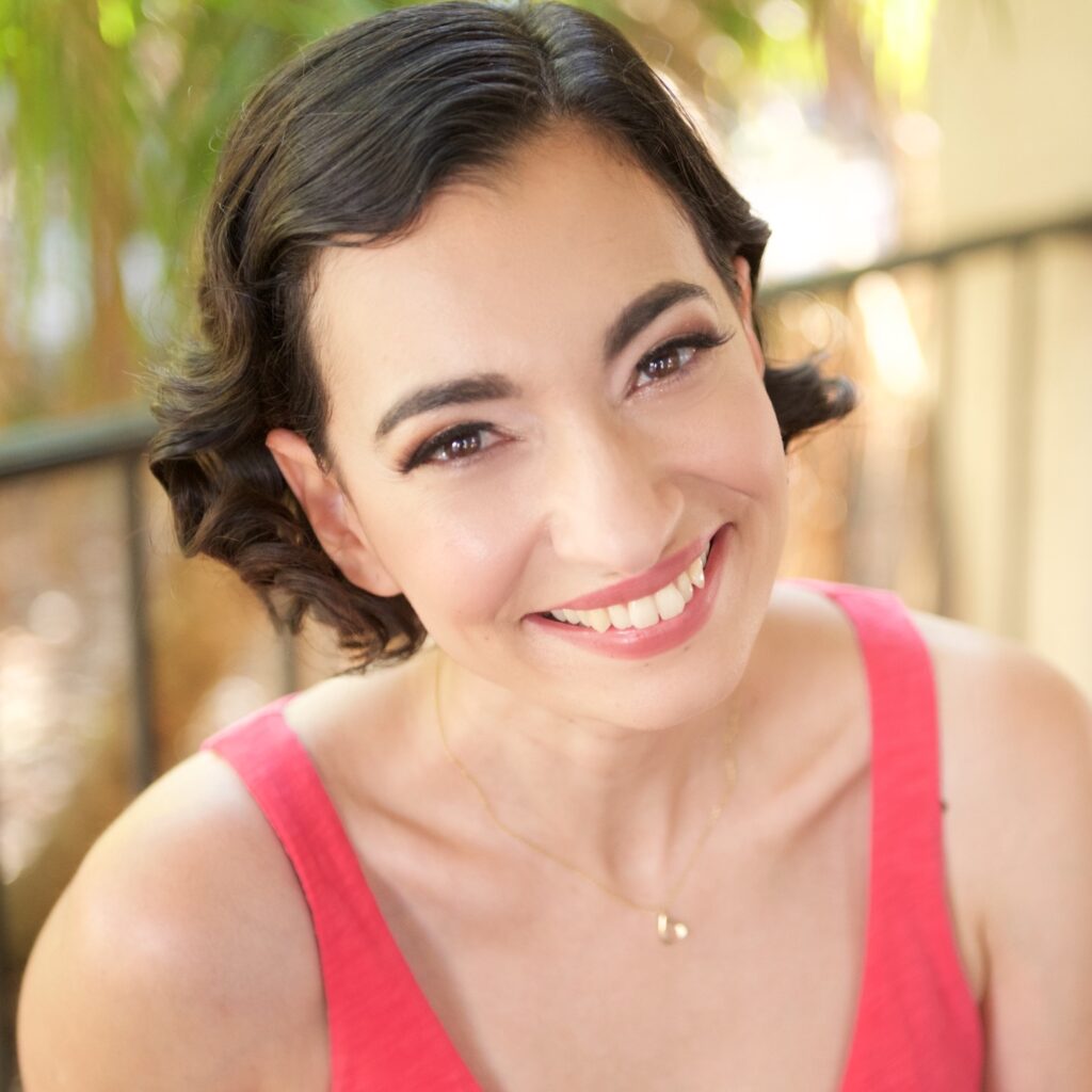 Headshot of a woman in her 30s who has heritage from Iran with light skin tone and chin length curly dark brown hair. She is wearing a coral tank top, gold necklace and smiling at the camera.