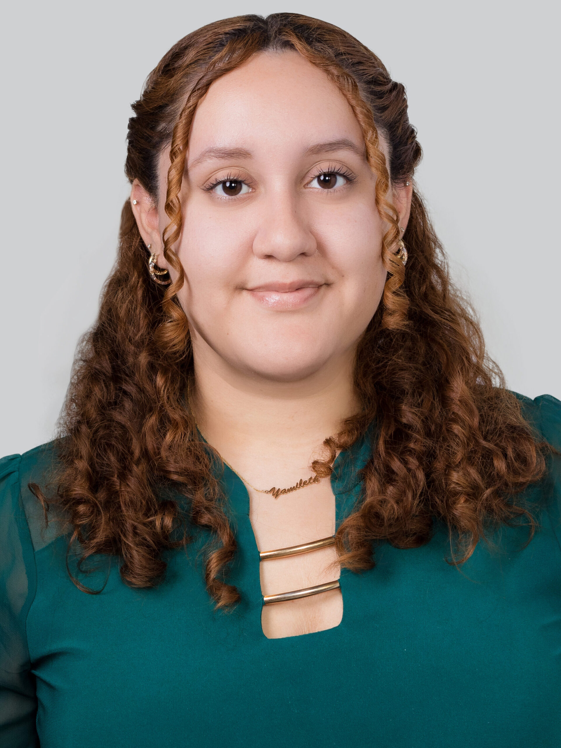 Headshot of a woman in her 20s who has light skin tone and curly dyed light red hair. She is wearing an emerald green blouse with gold accents, she has on a couple gold hoop earrings. She is smiling at the camera.