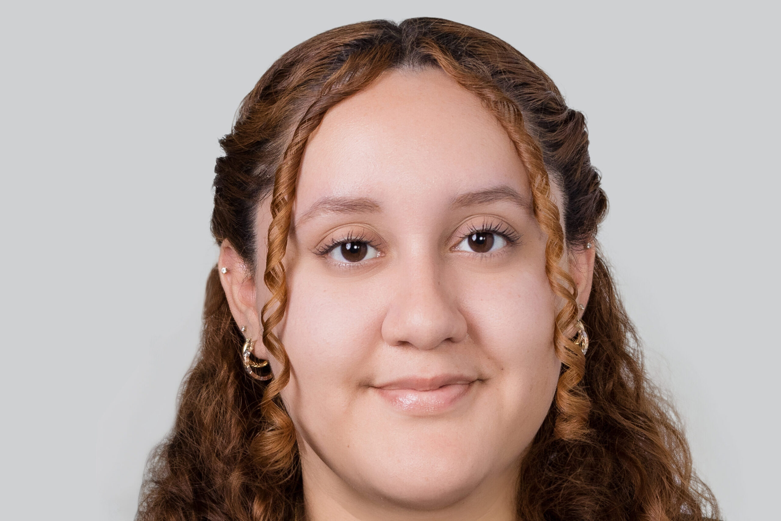 Headshot of a woman in her 20s who has light skin tone and curly dyed light red hair. The photo is too zoomed in to see what she is wearing, she has on a couple gold hoop earrings. She is smiling at the camera.