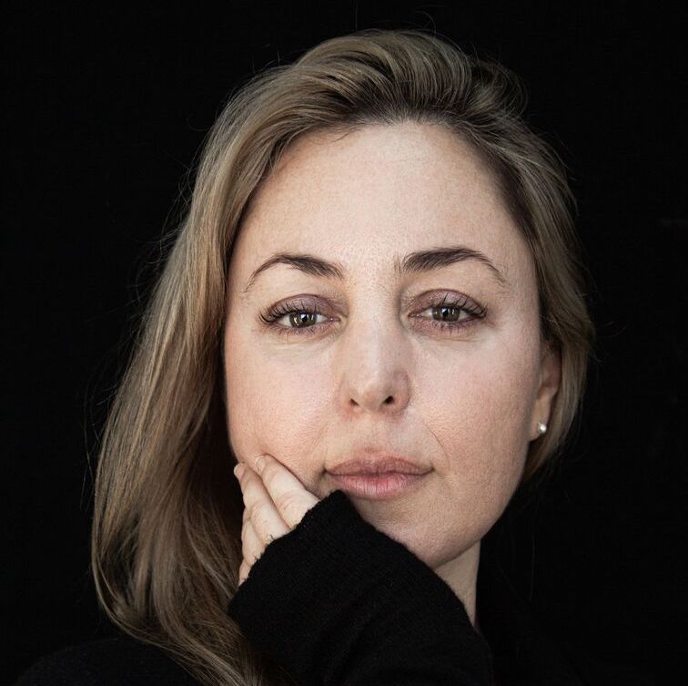 Headshot of a woman in her 30s who has heritage from Italy with light skin tone and straight shoulder length blonde hair. She is wearing a black sweater and pearl stud earrings, her right hand is resting on her chin, black sweater sleeve covering her hand. She is looking at the camera. 