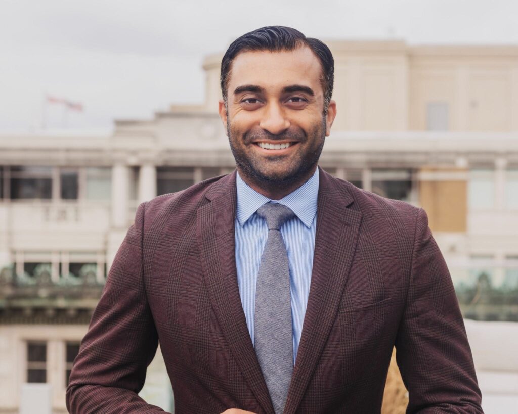 A photo of Akash smiling outside with a building in the distance. He is wearing a dark maroon suit jacket, a blue collared shirt and a tie.