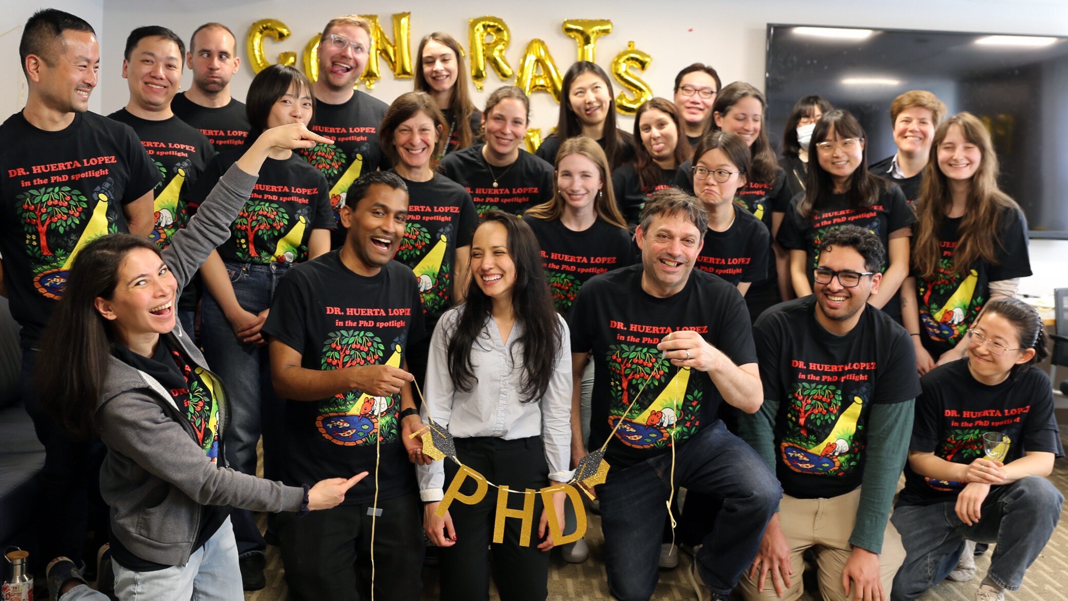 Silvia Huerta Lopez celebrates her PhD graduation with her lab mates in 2025. A large group of students surround Silvia, holding up a sign that says "PhD" in gold letters.