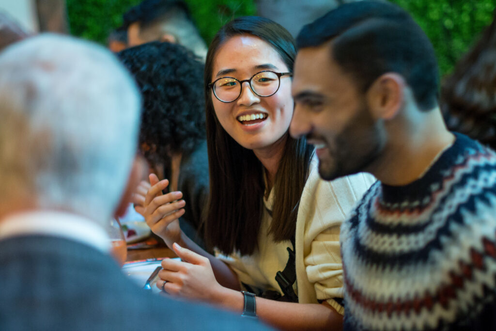 A candid photo of a dinner - Kathy Ku is at a table talking with a newer Fellow. They are engaged in conversation and you can see the back of another person's head in the photo.