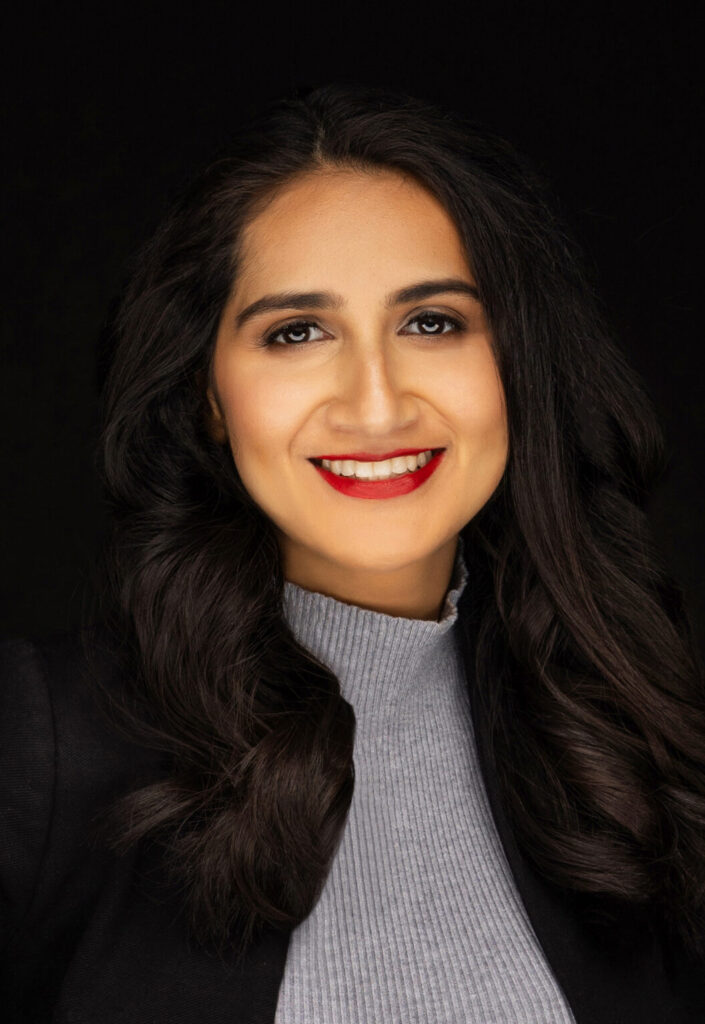 A photo of Riana Shah who is smiling and looking at the camera. She is wearing a grey turtleneck and a black coat and her hair is down.