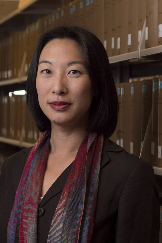A photo of Ming Hsu Chen who is wearing a maroon scarf and standing in a room with files behind her. She has a slight closed-lip smile and is posed for the camera. 
