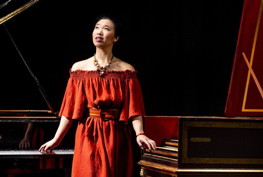 A photo of Hilda Huang is wearing a red dress and putting her hand on a large instrument.